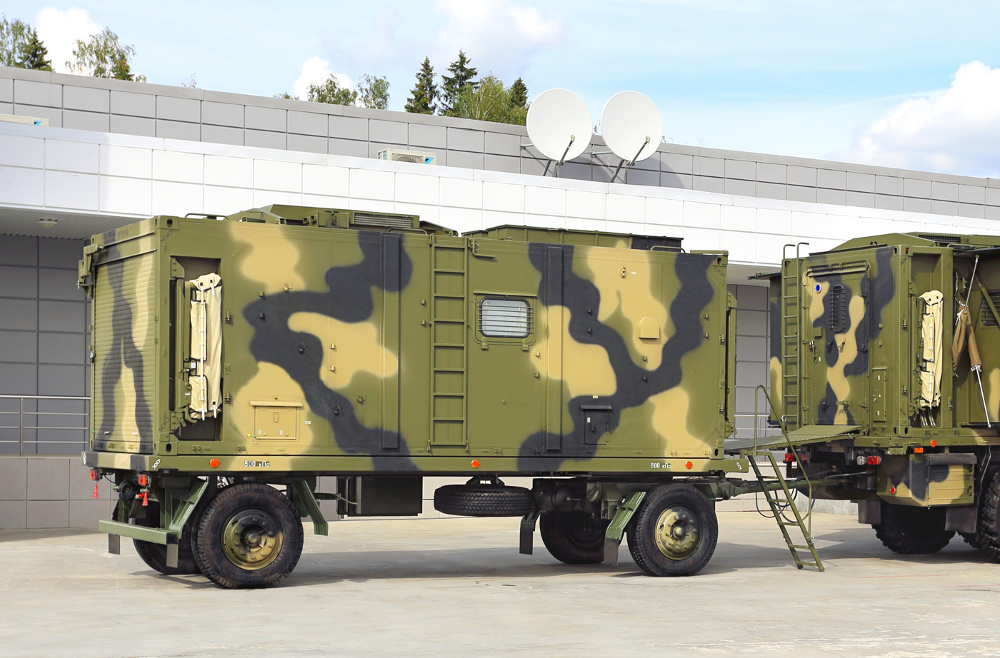 Two camo mobile military applications hitched together parked in front of a building